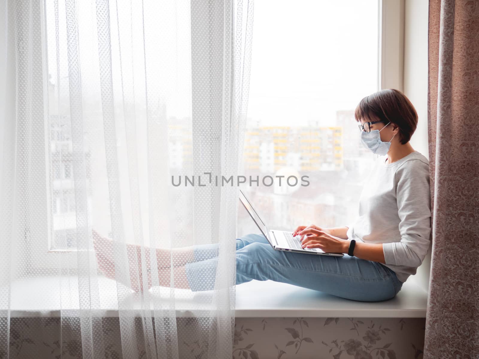 Woman in medical mask remote works from home. She sits on window by aksenovko