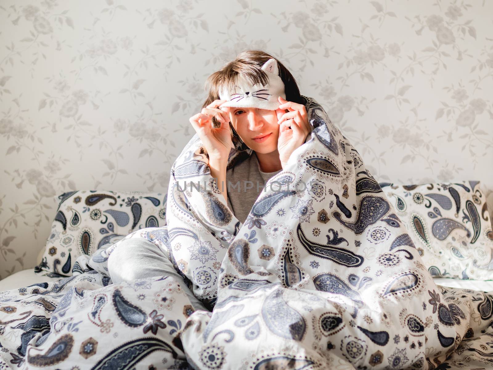 Young woman in grey pajama and sleeping mask in shape of cute sleeping cat face. She is just woke up and sit in bed. Early morning in cozy home.
