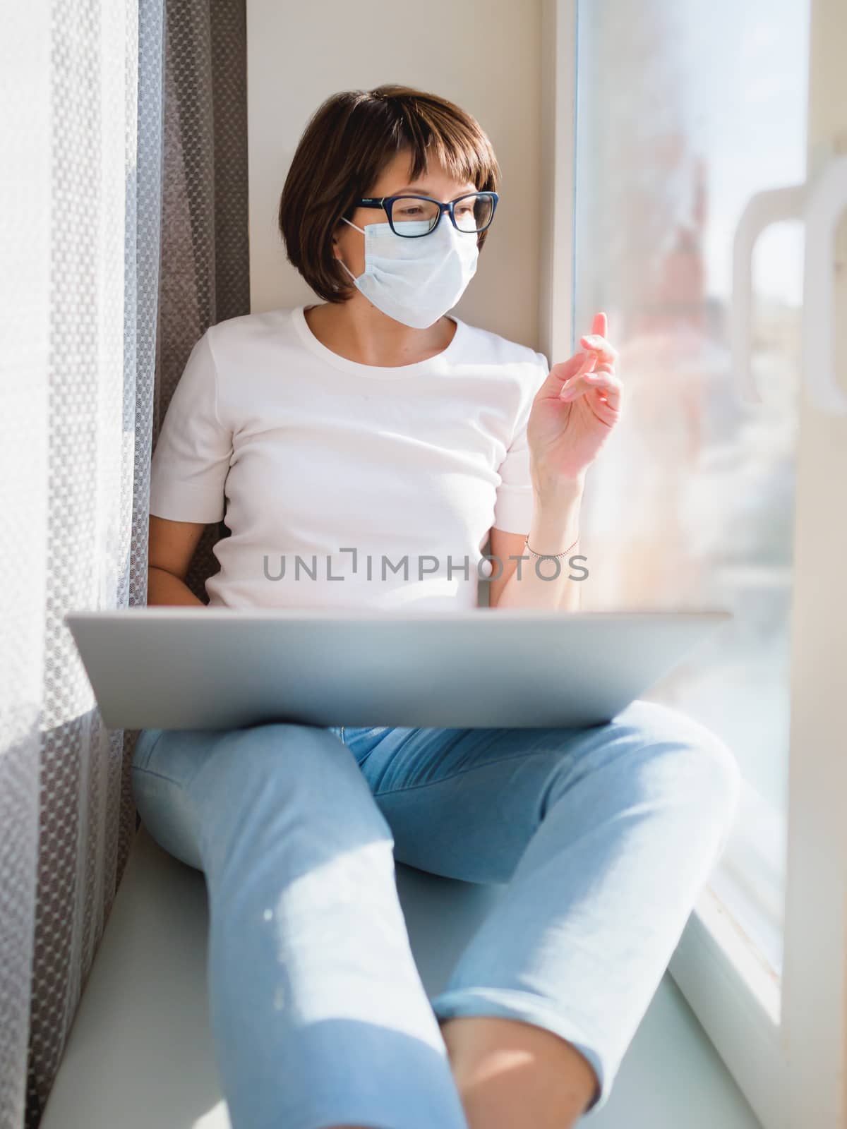 Woman in medical mask remote works from home. She sits on window sill with laptop on knees. Lockdown quarantine because of coronavirus COVID19. Self isolation at home.