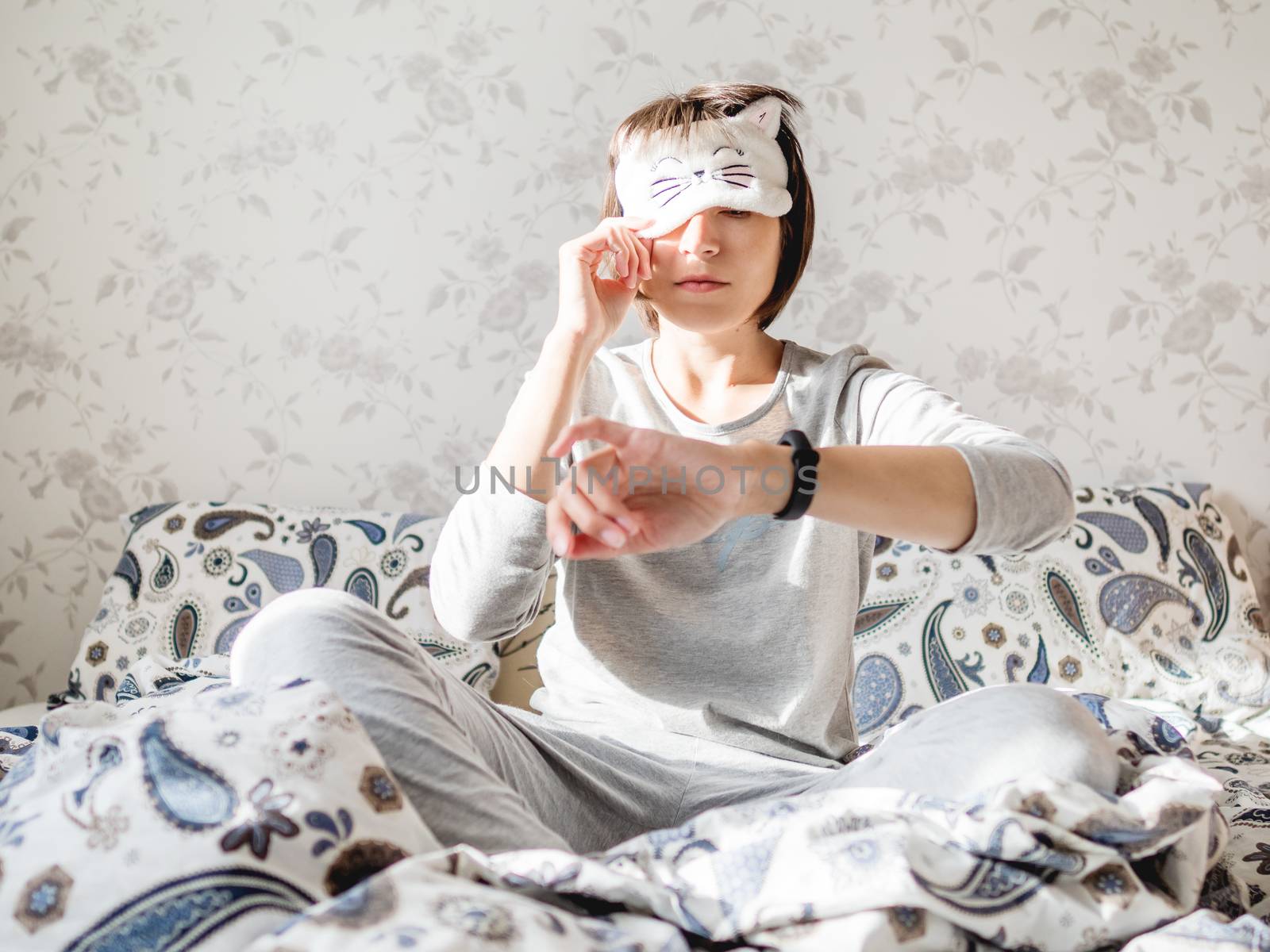 Young woman in grey pajama and sleeping mask in shape of cute sleeping cat face. She is just woke up and sit in bed. Early morning in cozy home.