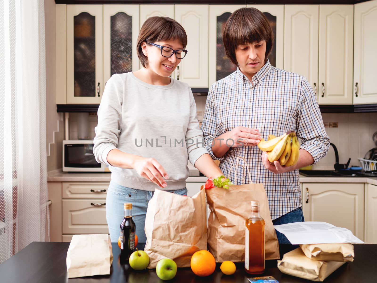 Young couple is sorting out purchases in the kitchen. Products in bags made of craft paper. Food delivery in conditions of quarantine because of coronavirus COVID19.