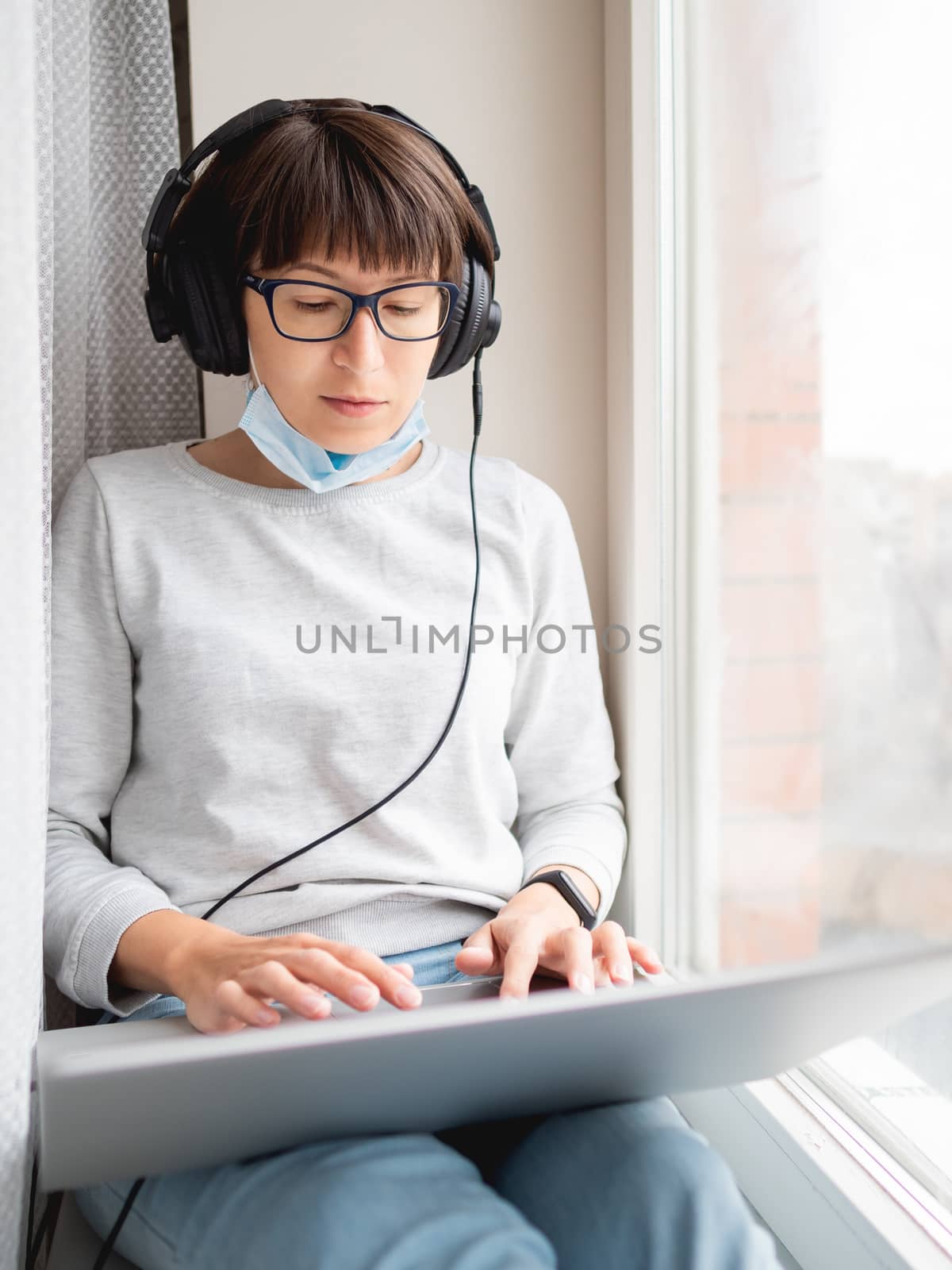 Woman in medical mask remote works from home. She sits on window sill with laptop on knees and headphones. Lockdown quarantine because of coronavirus COVID19. Self isolation at home.