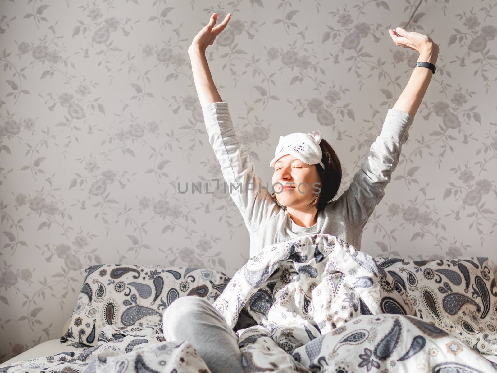 Young woman in grey pajama and sleeping mask in shape of cute sleeping cat face. She is just woke up and sit in bed. Early morning in cozy home.