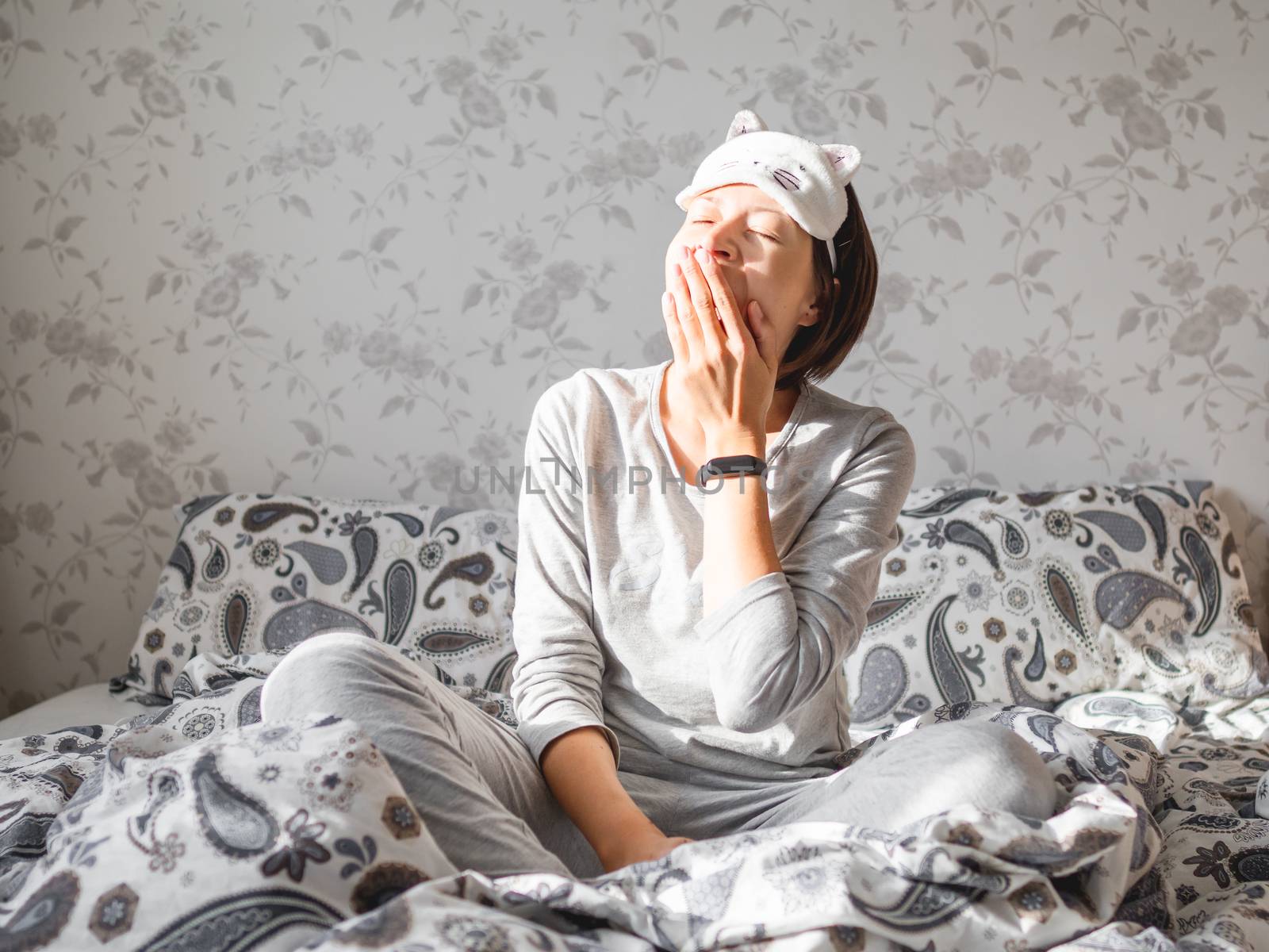 Young woman in grey pajama and sleeping mask in shape of cute sleeping cat face. She is just woke up and sit in bed. Early morning in cozy home.