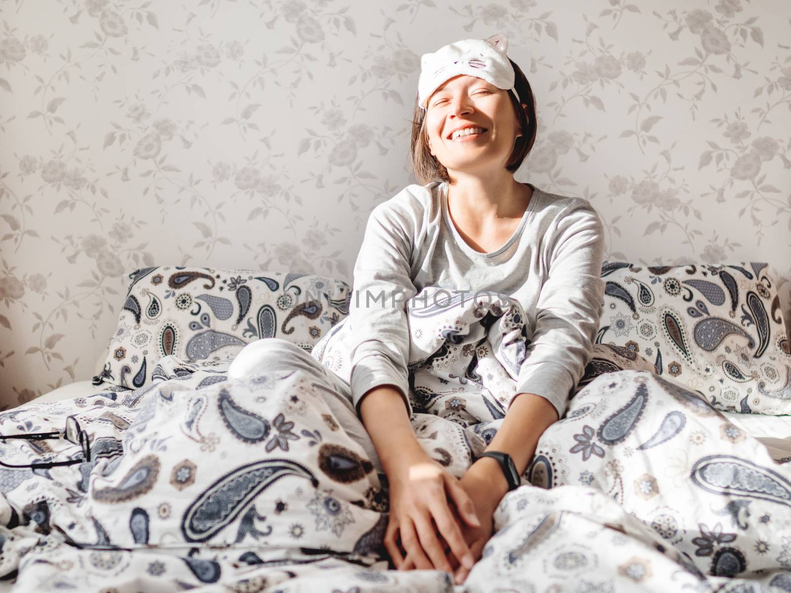 Young woman in grey pajama and sleeping mask in shape of cute sleeping cat face. She is just woke up and sit in bed. Early morning in cozy home.