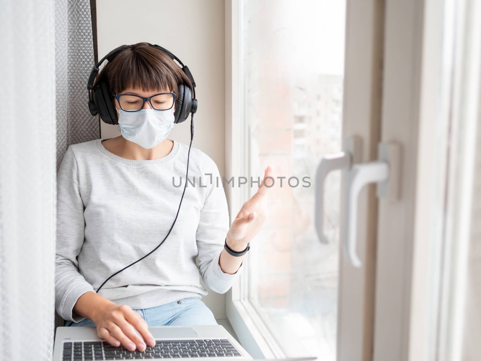 Woman in medical mask remote works from home. She sits on window sill with laptop on knees and headphones. Lockdown quarantine because of coronavirus COVID19. Self isolation at home.