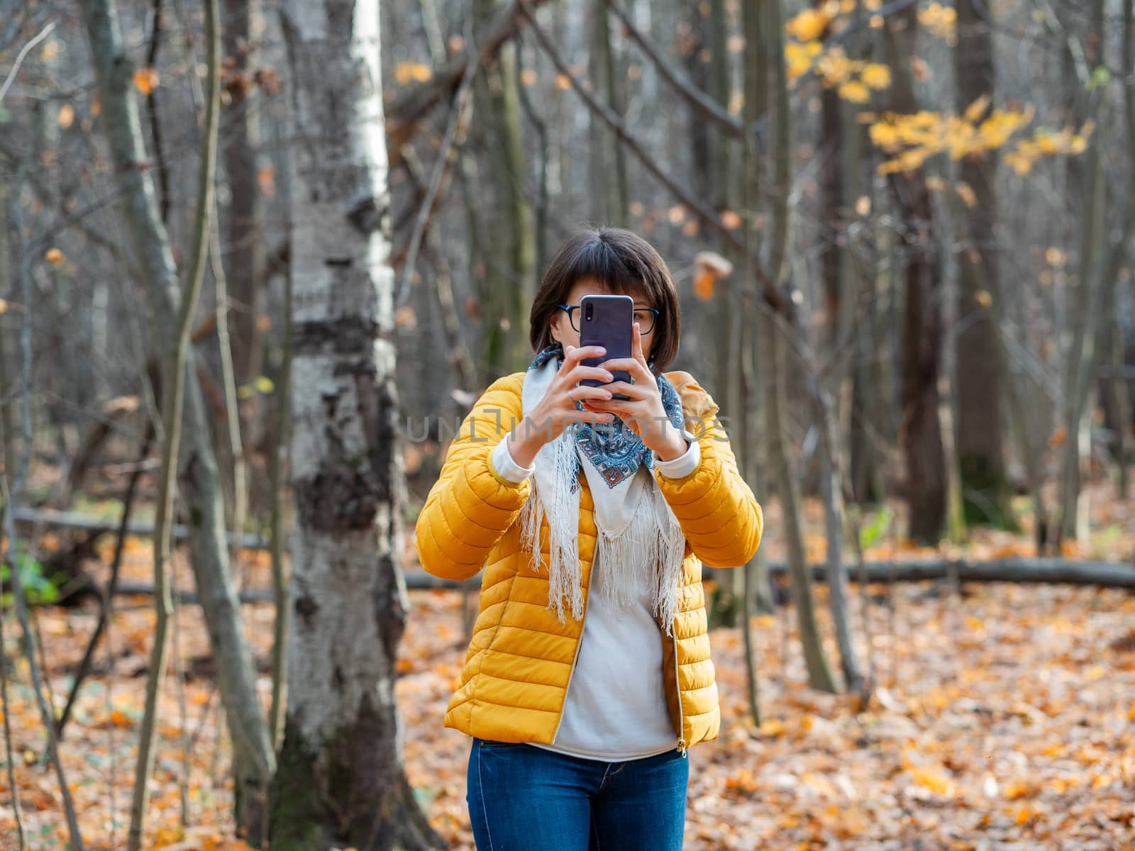 Woman in bright yellow jacket with smartphone. Young girl with wide smile walking and taking pictures of autumn forest.