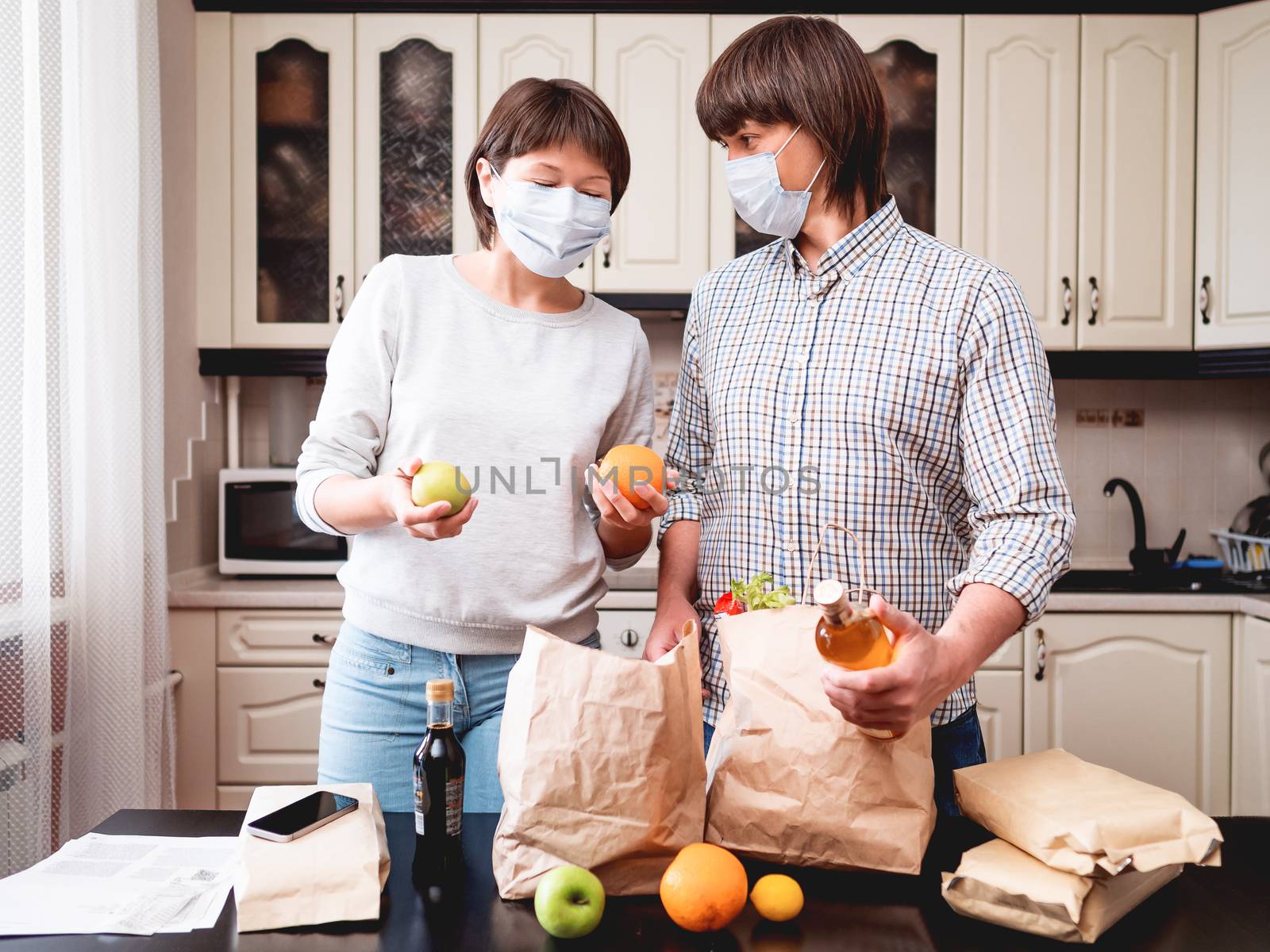 Young couple in protective masks is sorting out purchases in the kitchen. Products in bags made of craft paper. Food delivery in conditions of quarantine because of coronavirus COVID19.