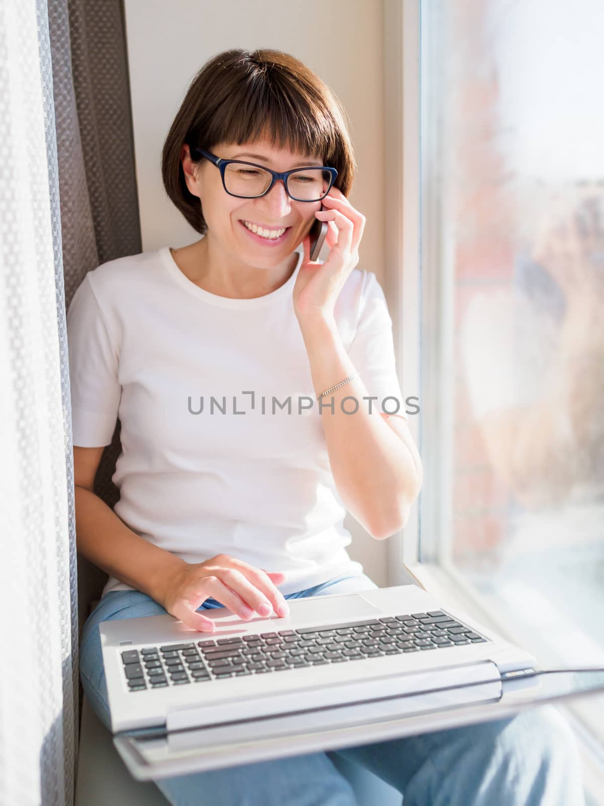Woman with smartphone remote works from home. She smiles widely on window sill with laptop on knees. Lockdown quarantine because of coronavirus COVID19. Self isolation at home.