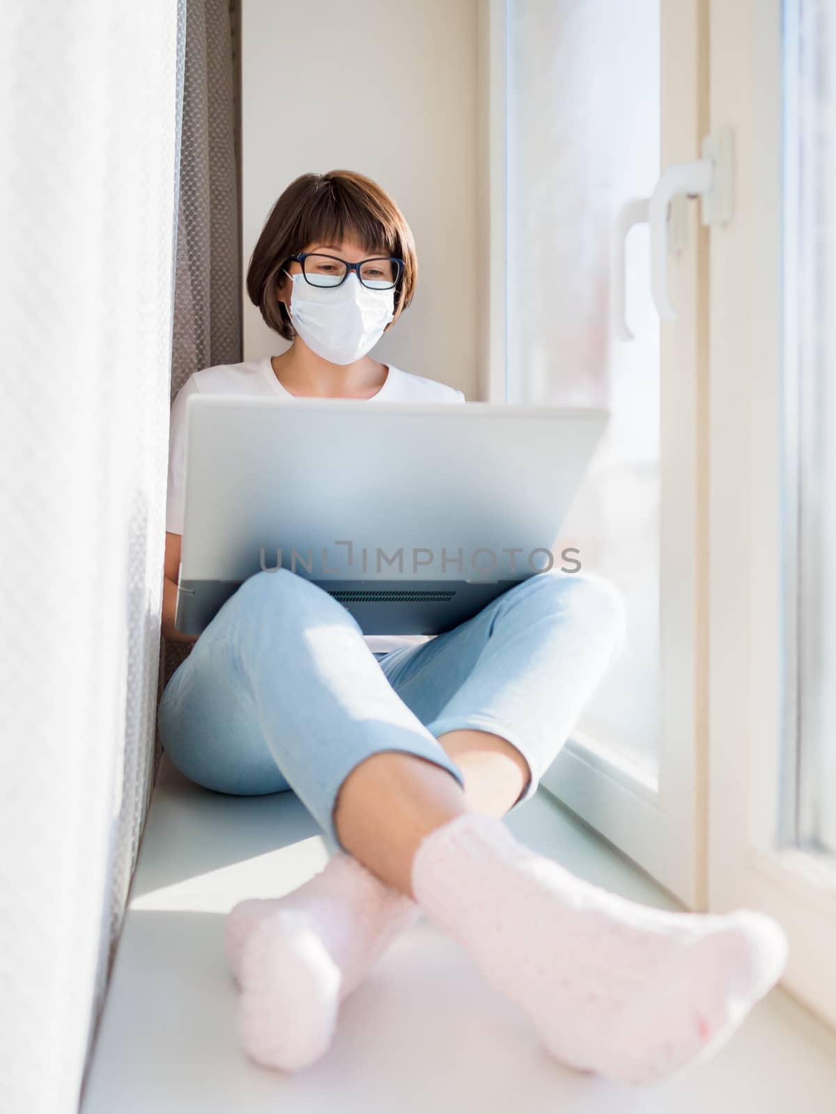 Woman in medical mask remote works from home. She sits on window sill with laptop on knees. Lockdown quarantine because of coronavirus COVID19. Self isolation at home.