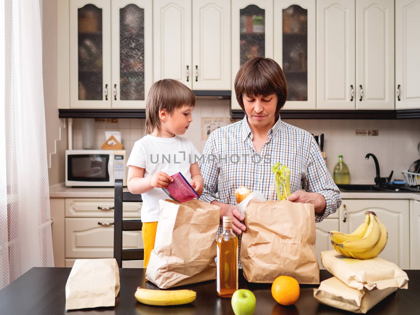 Family sorts out purchases in the kitchen. Father and son tastes products in bags made of craft paper. Food delivery in conditions of quarantine because of coronavirus COVID19.