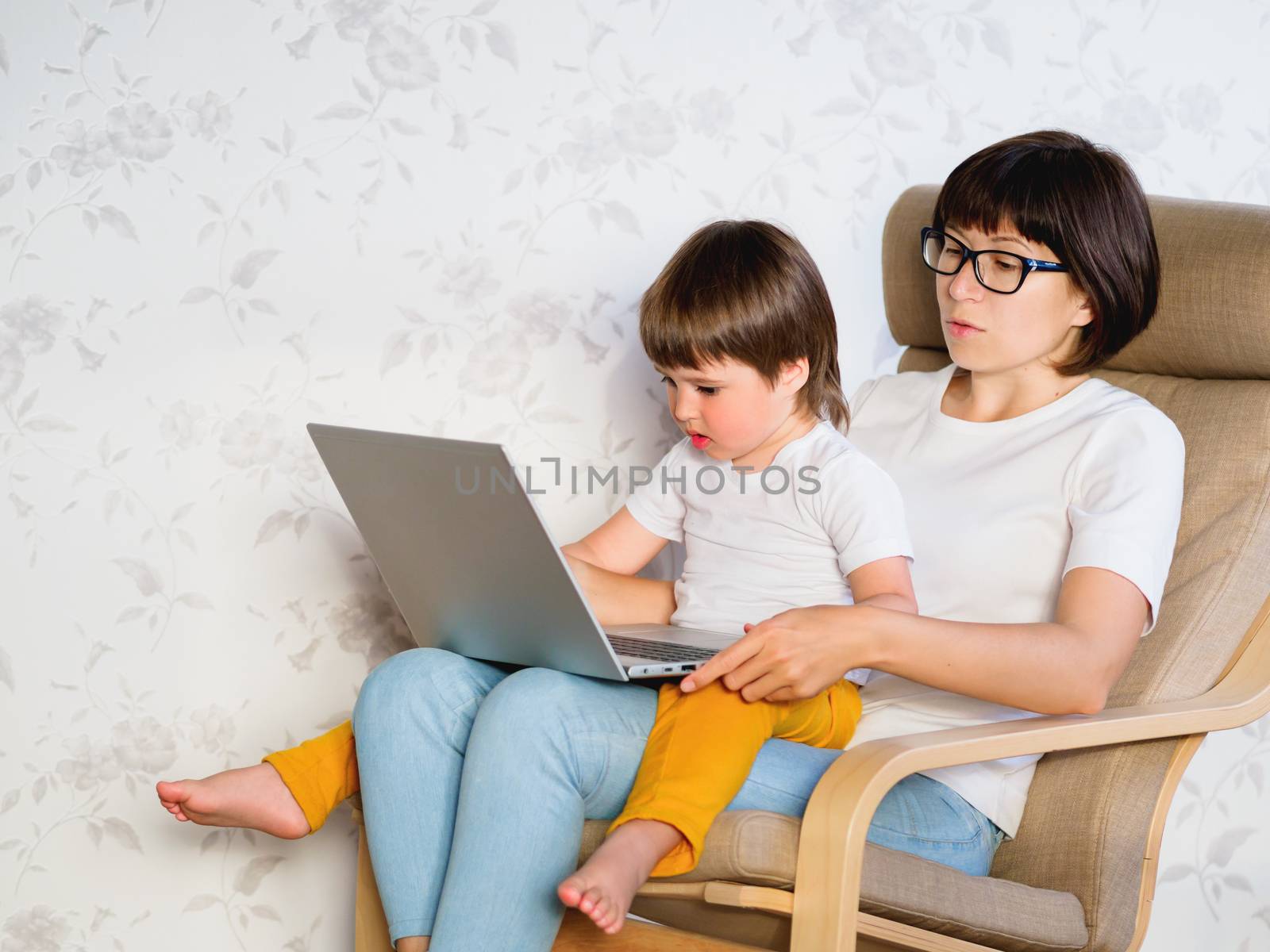 Mother and her toddler boy sit together on chair with laptop. Wo by aksenovko