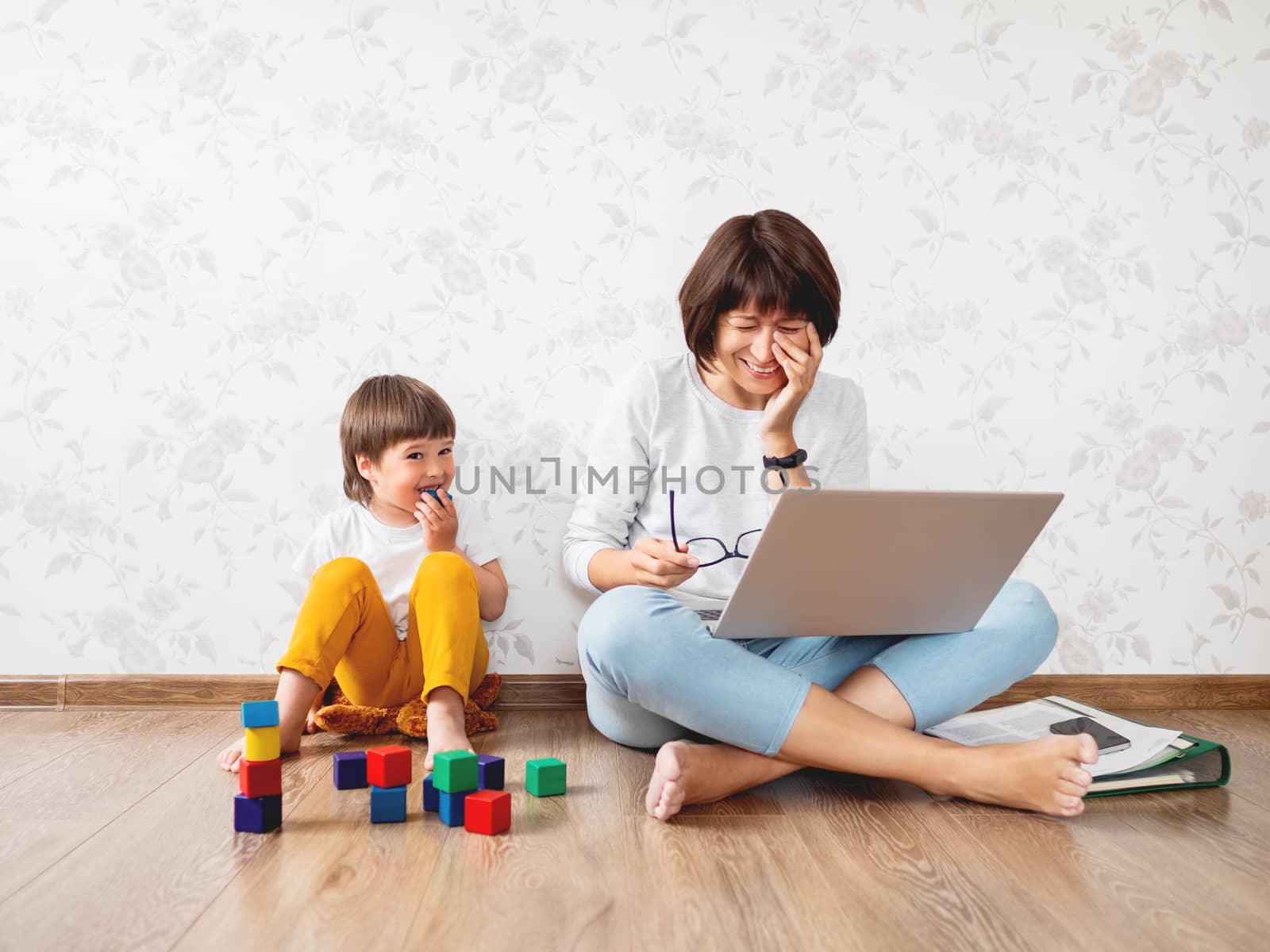 Family sits at home quarantine because of coronavirus COVID19. Mother remote works with laptop, son plays with toy blocks. Self isolation at home.