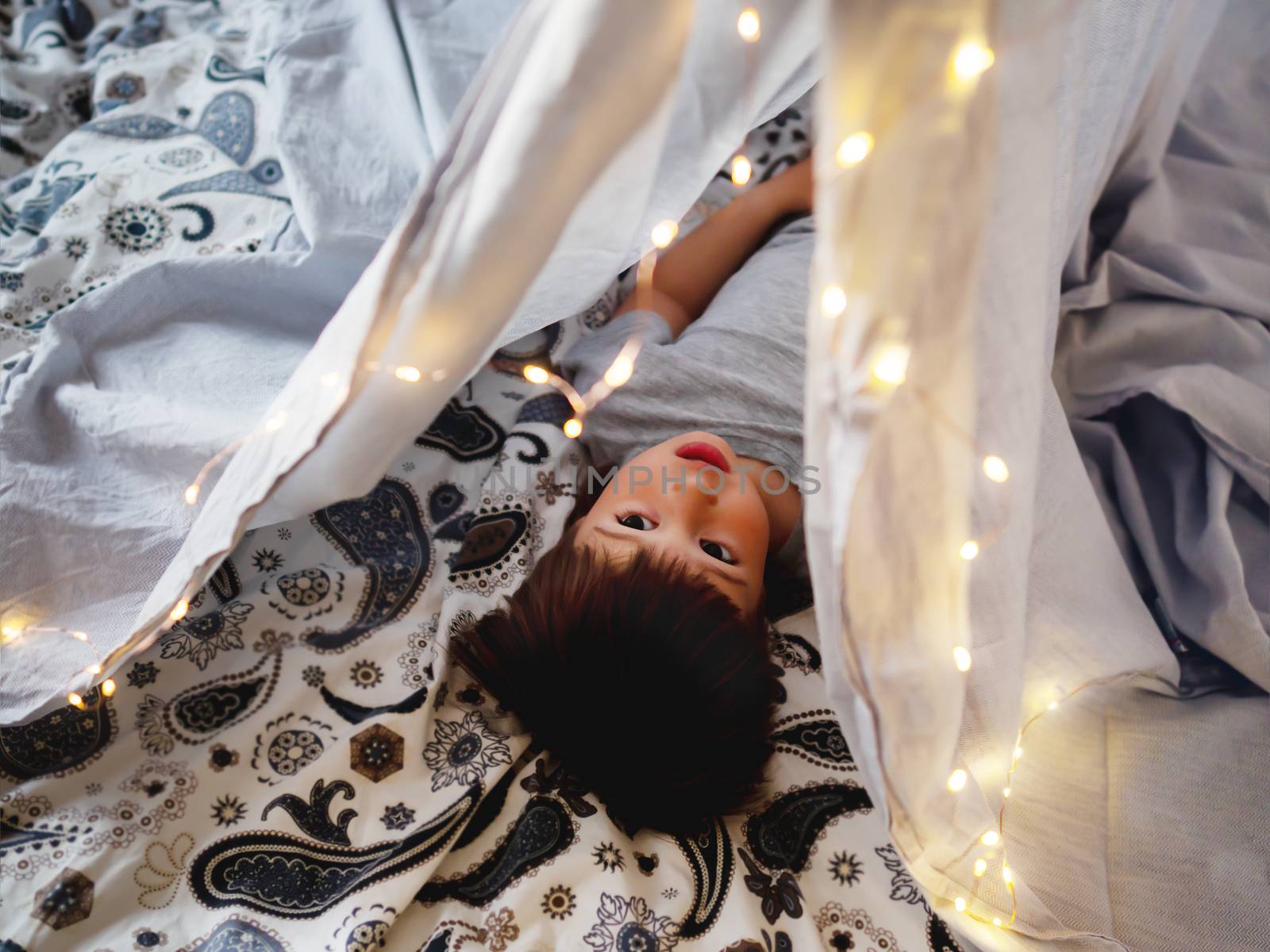 Little boy lays inside DIY tent in bedroom. Toddler plays in tent made of linen sheet on bed. Cozy evening at home.