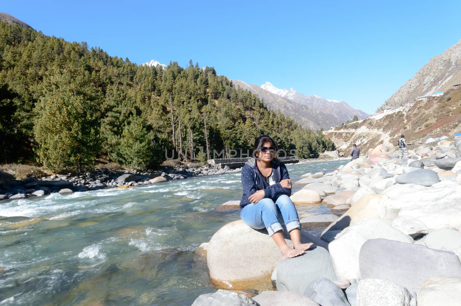 Front View of Solo Indian traveler in winter casual sitting alone near stream flowing water of remote mountain valley. Snow capped Himalayan mountain forest blue sky in background. Spiti Valley Himachal Pradesh India South Asia Pac by sudiptabhowmick