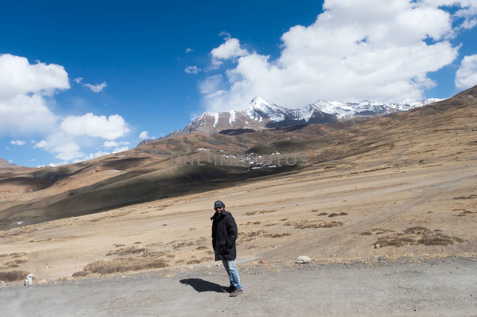Solo Indian traveler in winter clothing standing alone in a remote mountain road. Snow capped mountain with floating clouds blue sky in the background.
