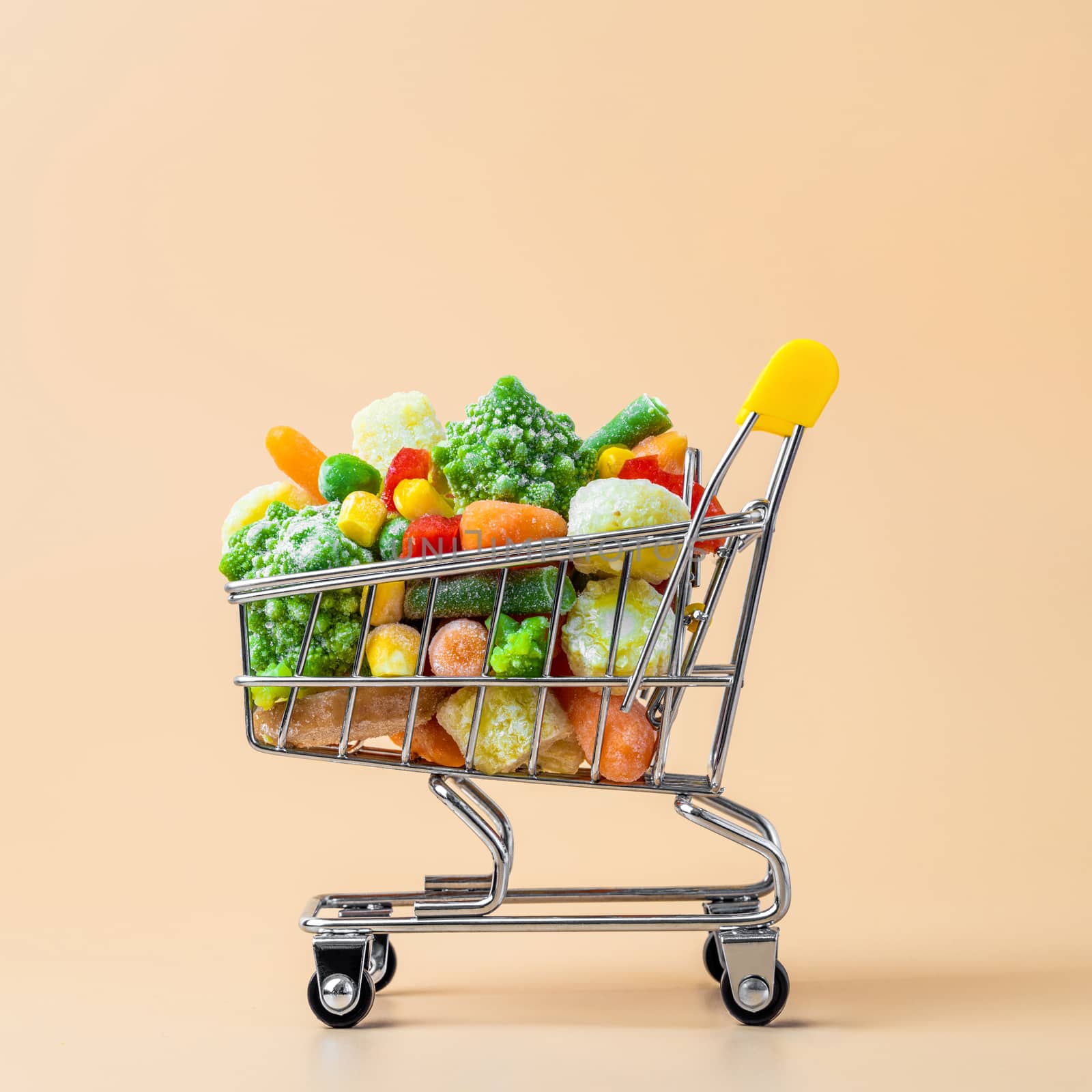 Frozen vegetables assorted in toy shopping cart on cream background. Full of assorted frozen vegetables food shop trolley at beige or yellow backdrop. Minimalistic concept.