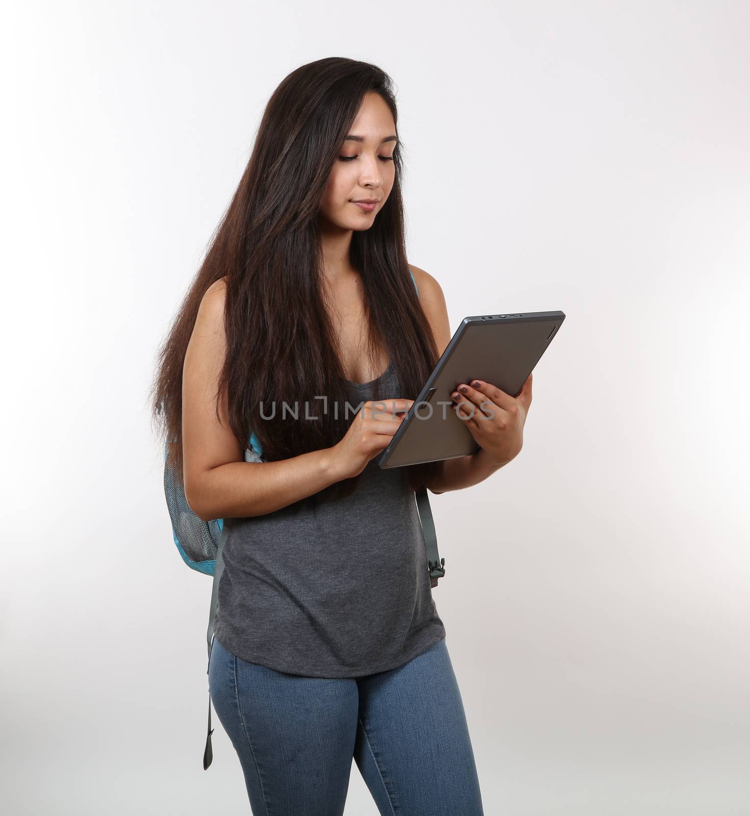 A young female student in jeans views her tablet as she holds her backpack.