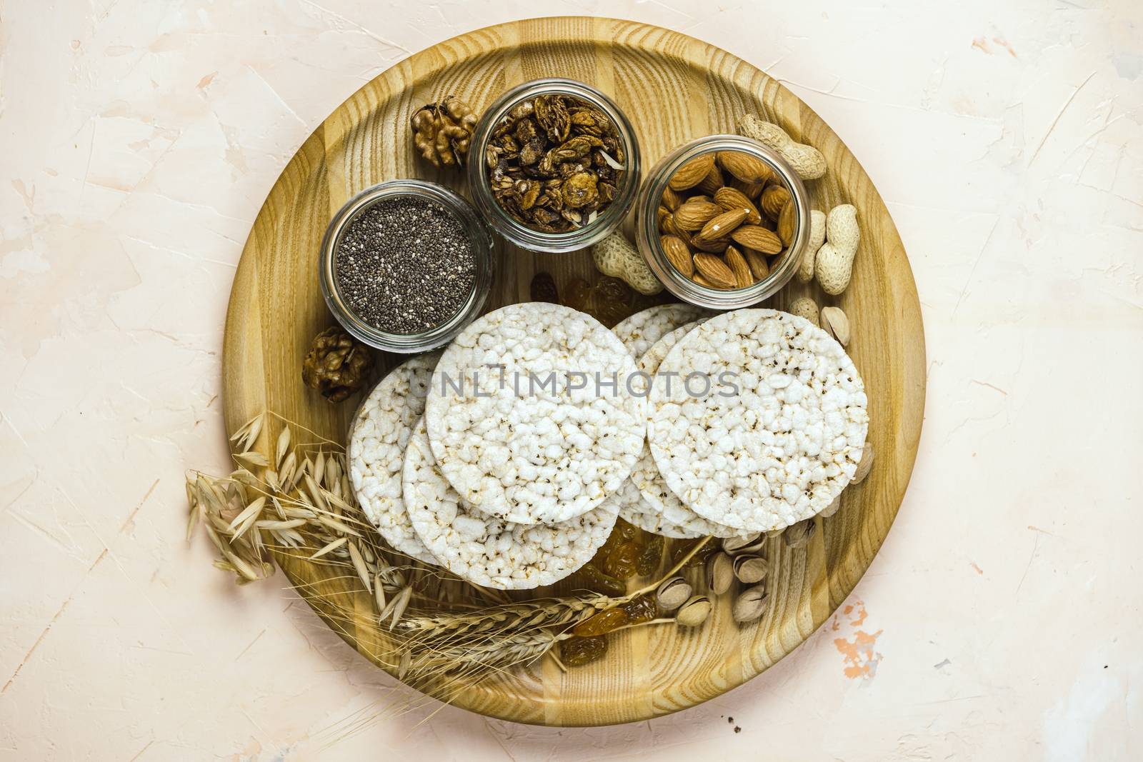 American puffed rice cakes. Healthy snacks with ears of wheat on round cutting wooden board on light pink concrete surface, top view, flat lay, copy space for you text	