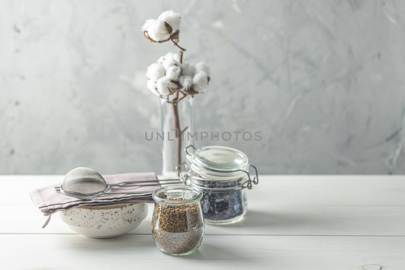 Coffee beans and instant coffee in glass jars, cotton flowers and kitchenware on white wooden table with grey concrete wall at background. Details of still life in the home interior. Cosy concept.