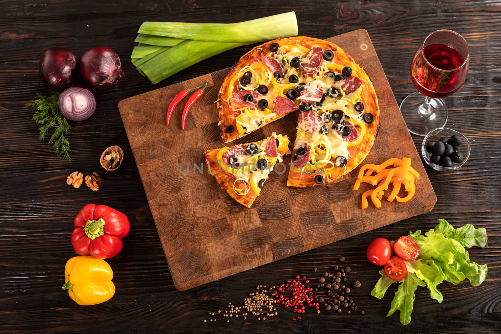 Pizza on a wooden cutting board next to vegetables, spices and a glass of wine