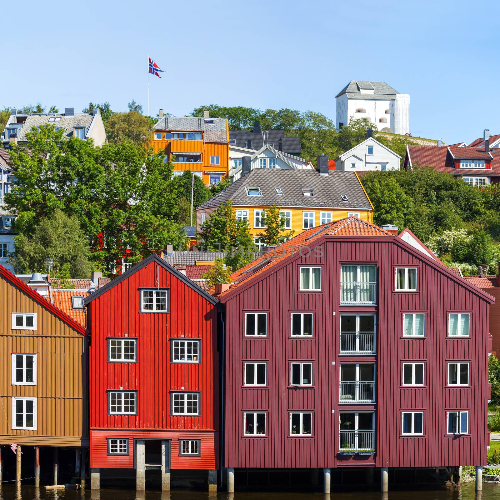 Famous wooden colored houses in Trondheim city, Norway. Colorful houses on stilts and flag of Norway in sunny day.