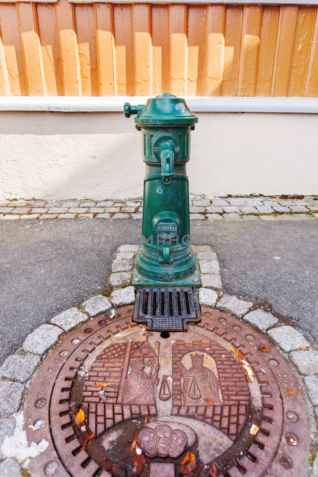 Metal sewerage hatchway and old fashioned water standpipe with bas relief. Iron manhole with historic scene. Trondheim, Norway.