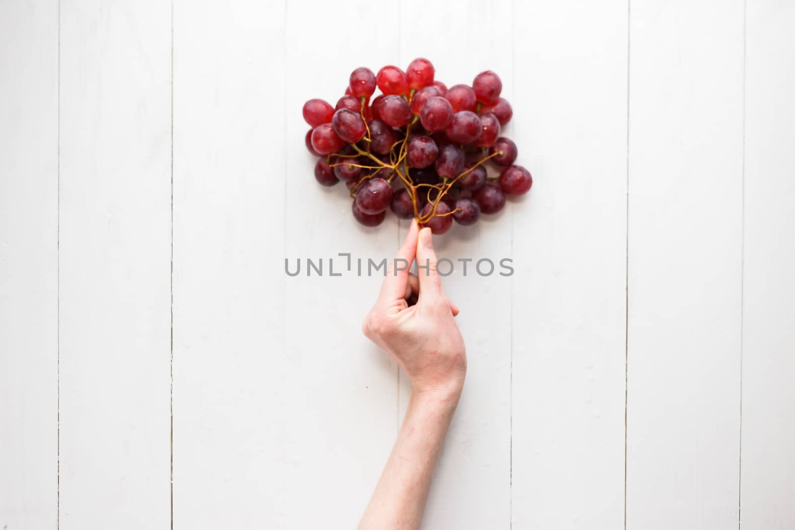 The girl's hand holds on to a bunch of red grapes on a wooden background. View from above. Grapes are like balloons. by vladdeep