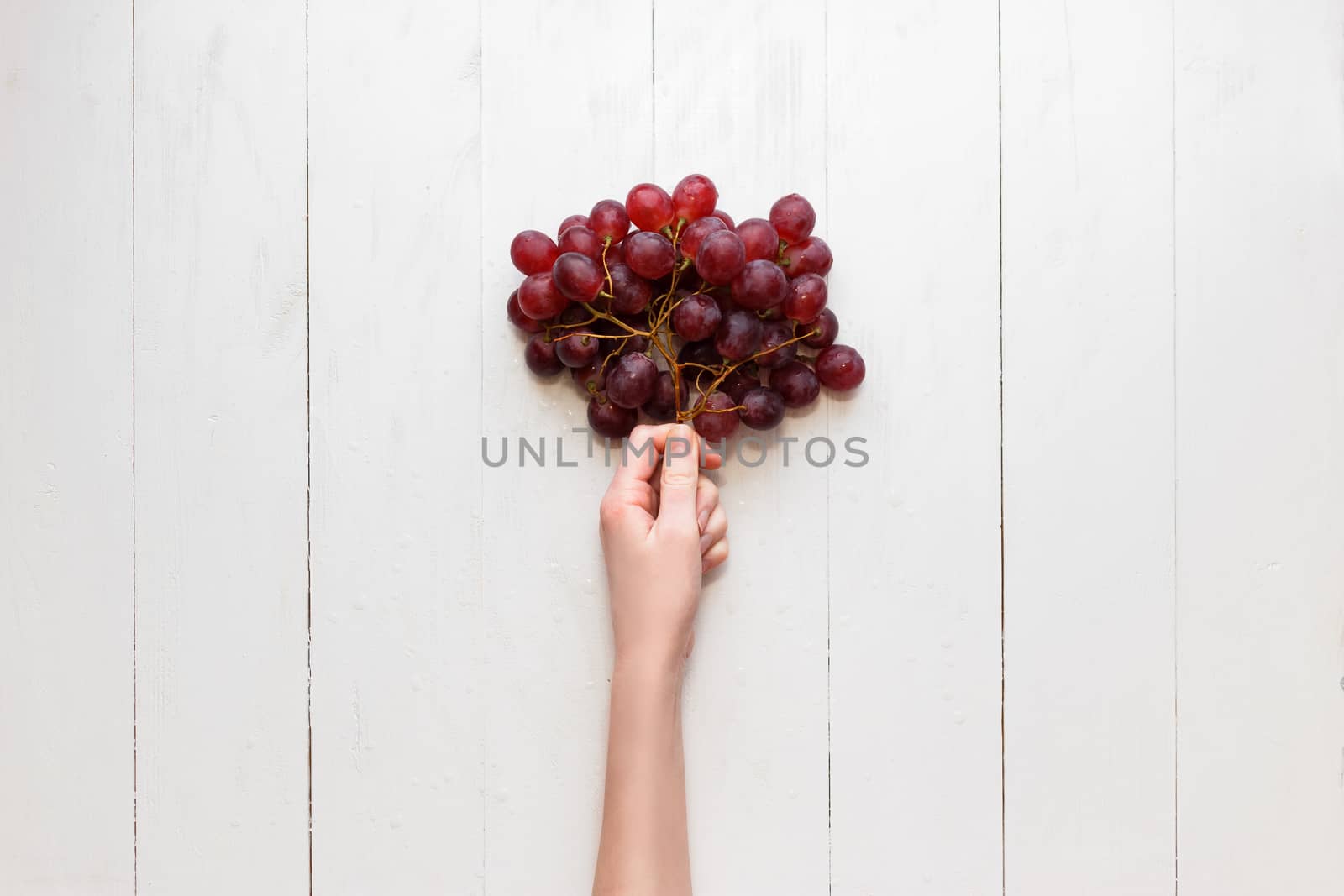 The girl's hand holds on to a bunch of red grapes on a wooden background. View from above. Grapes are like balloons. by vladdeep