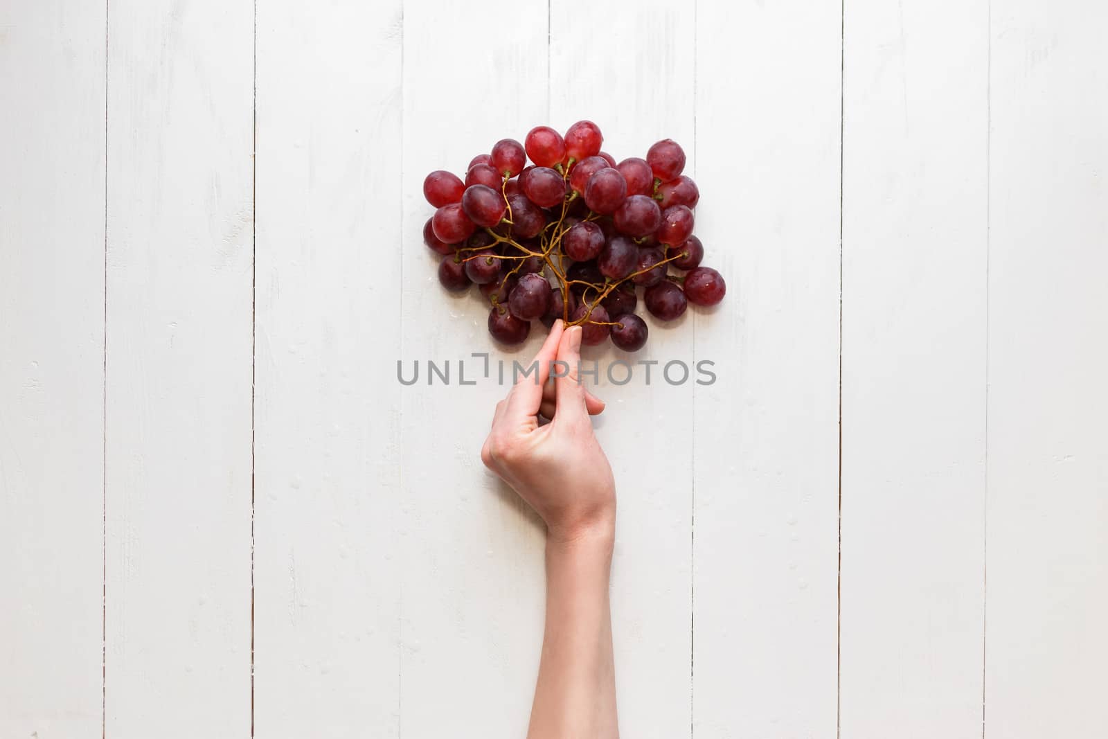 The girl's hand holds on to a bunch of red grapes on a wooden background. View from above. Grapes are like balloons. by vladdeep