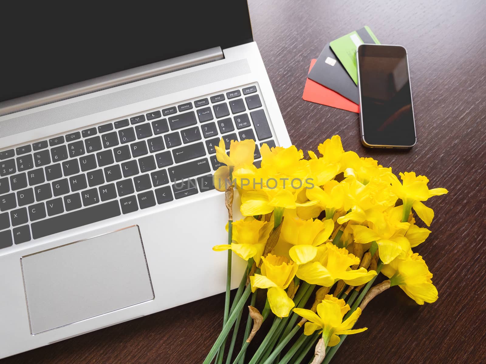 Bouquet of Narcissus or daffodils lying on silver metal laptop. Top view on bright yellow flowers on portable device and smartphone with credit cards on wooden table.