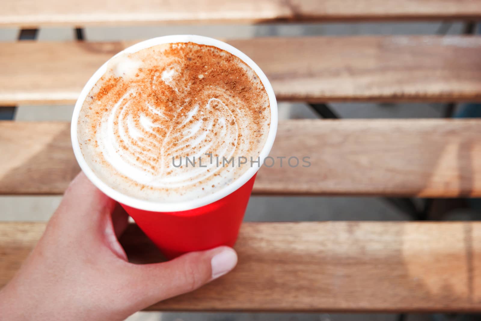 Woman holding a red paper cup with coffee. Coffee to go. Tasty hot beverage on wooden table in sunny day. Outdoors meal.