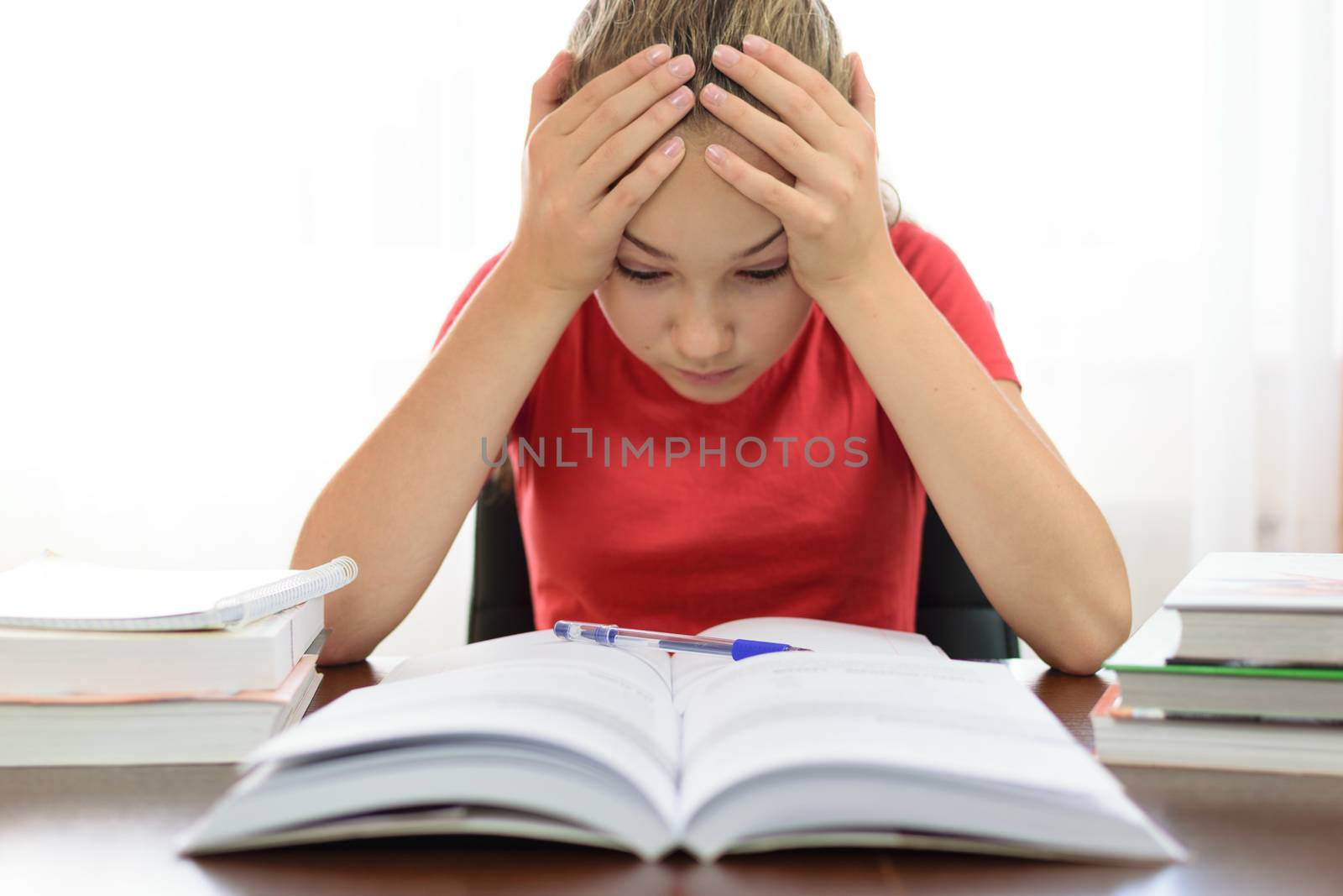 Schoolgirl holds her head in her hands over a pile of books on the desk and is tired and frustrated with the homework problem at school.