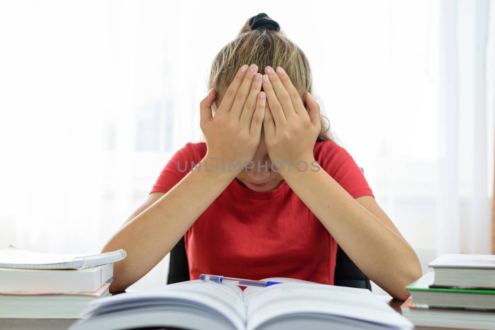 Schoolgirl holds her head in her hands over a pile of books on the desk and is tired and frustrated with problems at school.