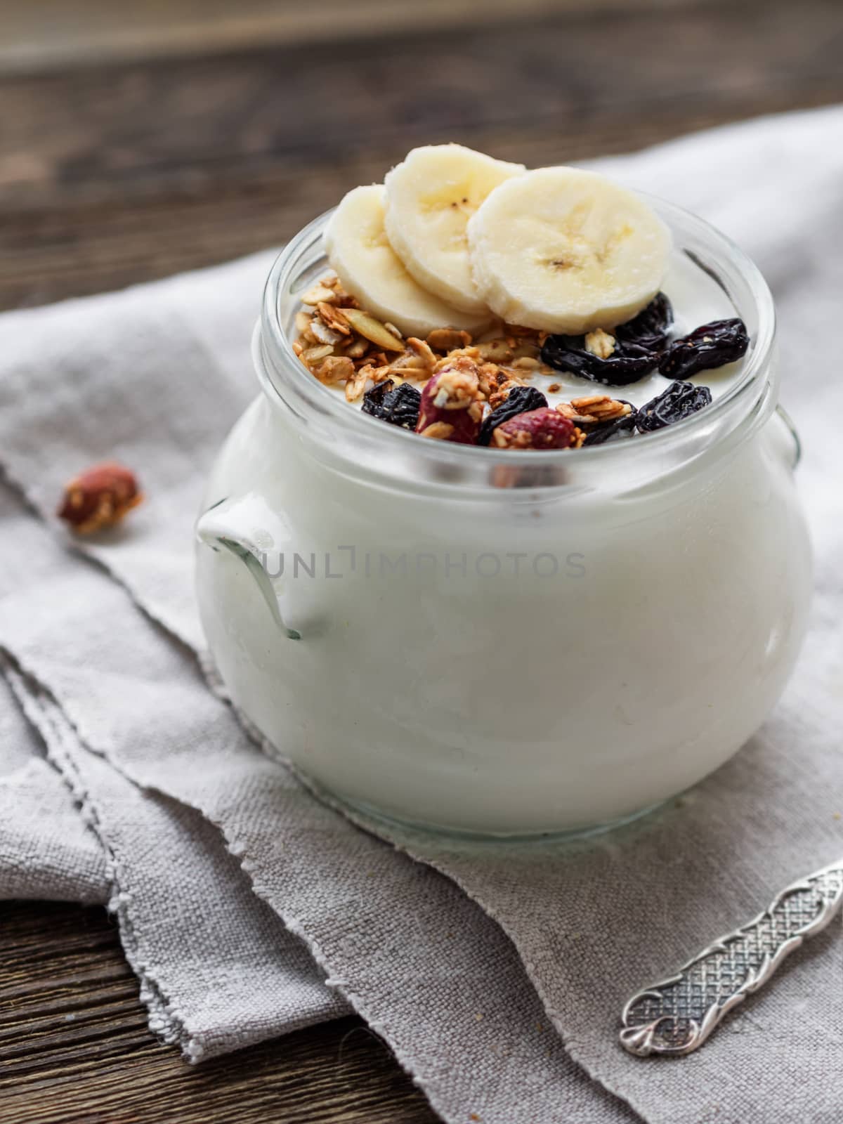 Natural homemade yogurt in a glass jar. Healthy food for breakfast with muesli. Jar with granola and banana slices on linen tablecloth on on wooden table.