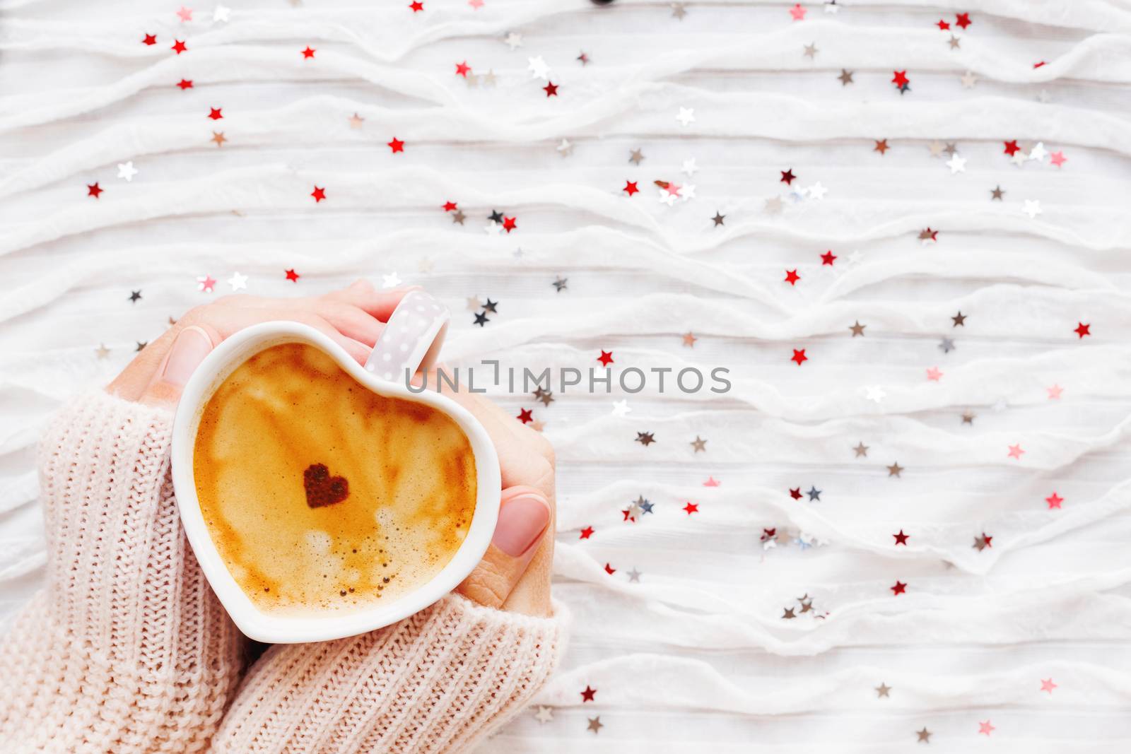 Woman holds a cup of hot coffee with cinnamon heart. Winter and Valentine's Day fabric background with sparkling silver and red confetti.