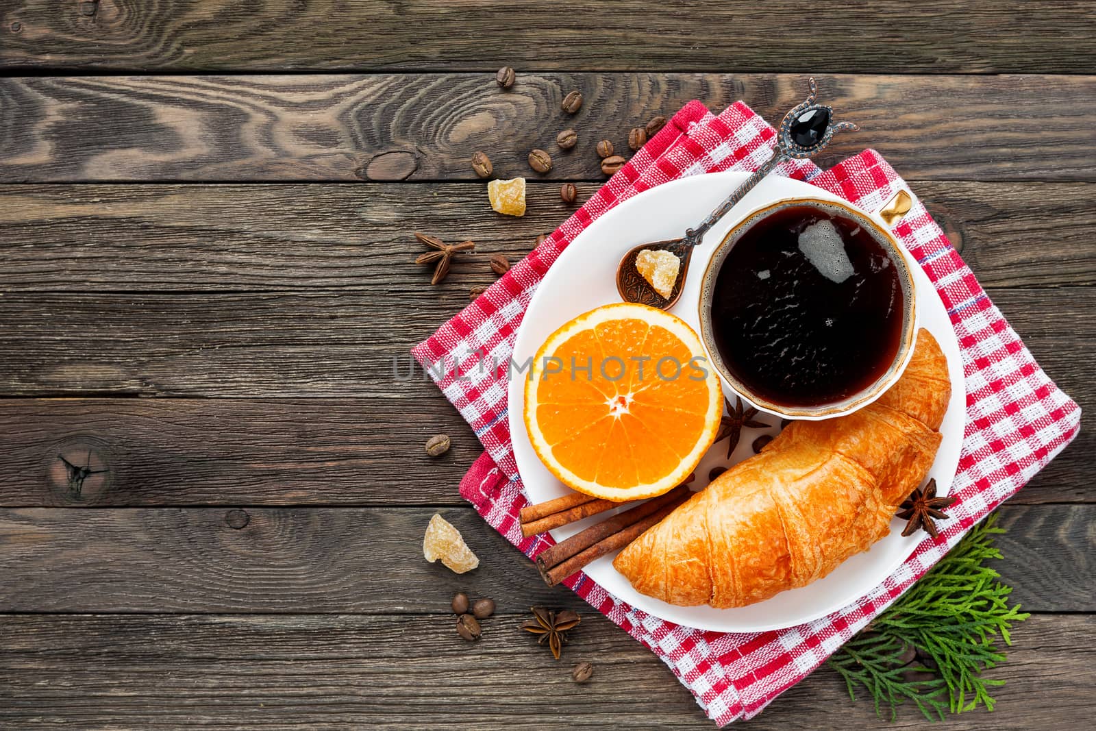Continental breakfast - cup of hot coffee, croissant and orange. Tasty food on plaid red napkin. Rustic wooden background. Top view, place for text.