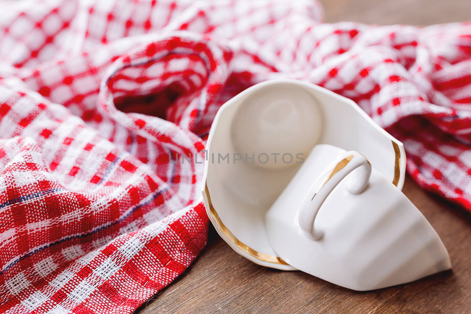 Broken white cup on wooden background with plaid red towel. Damaged mug with golden decoration.