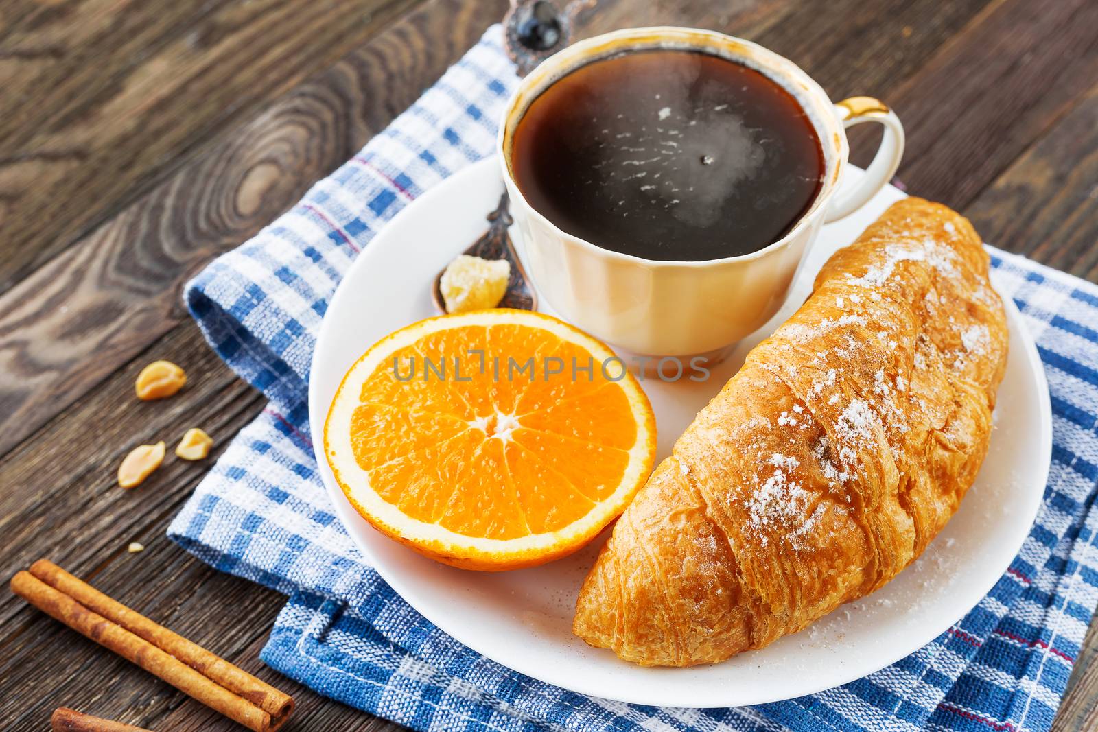 Continental breakfast - cup of hot coffee, croissant and orange. Tasty food on plaid blue napkin, rustic wooden background.