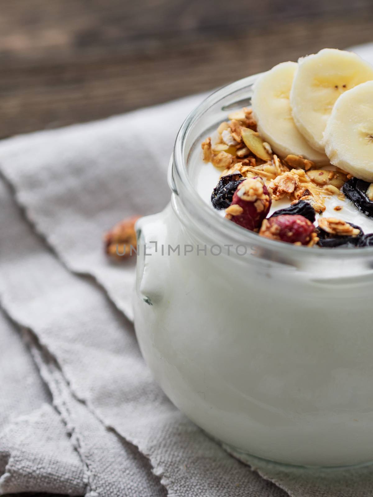 Natural homemade yogurt in a glass jar. Healthy food for breakfast with muesli. Jar with granola and banana slices on linen tablecloth on on wooden table.