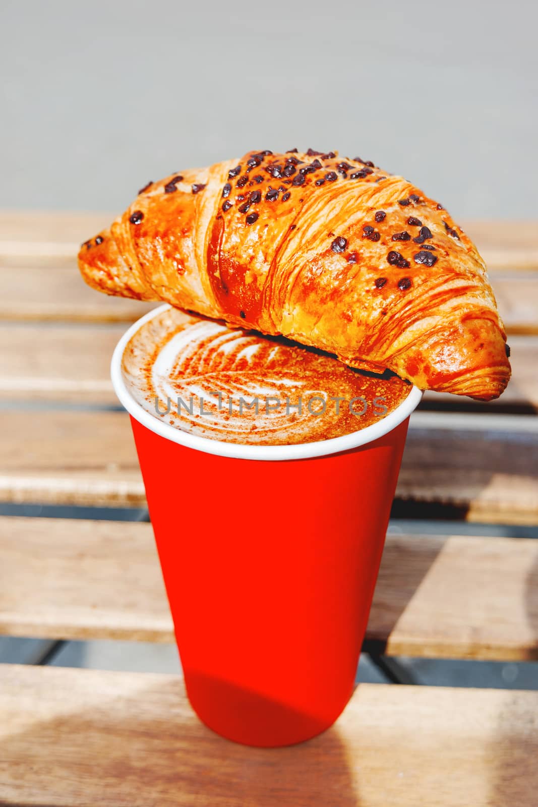 Red paper cup with coffee and chocolate croissant. Coffee to go. Tasty hot beverage on wooden table in sunny day. Outdoors meal.
