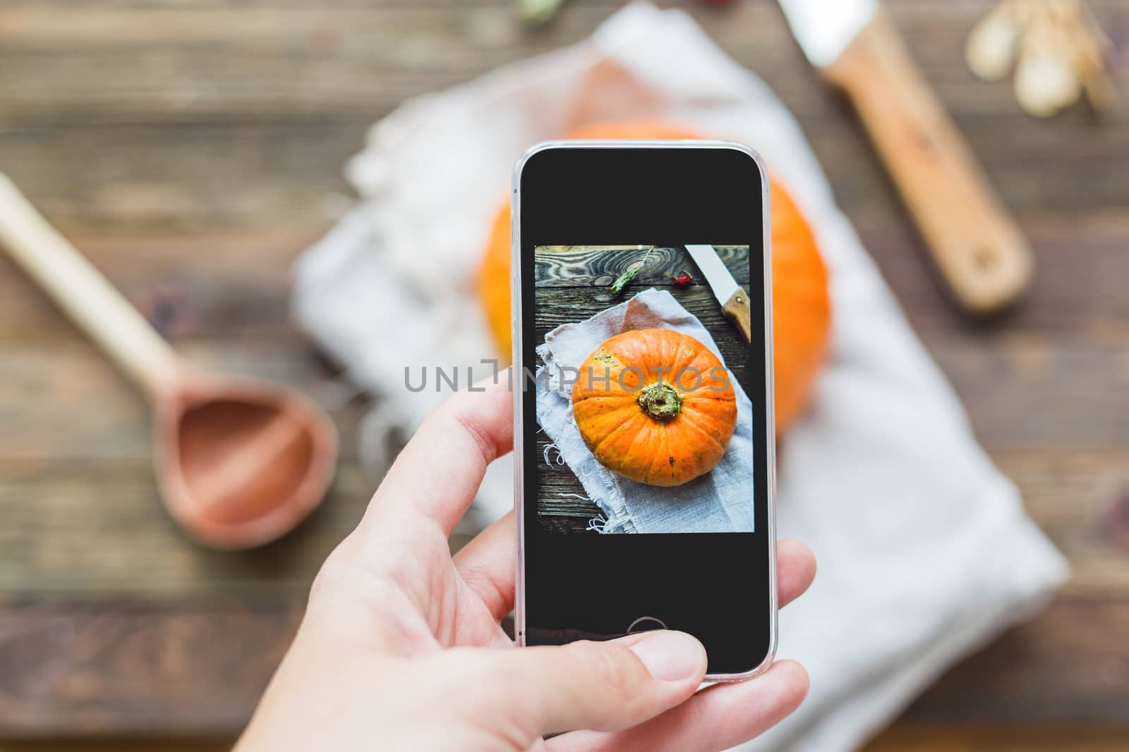 Woman shooting fresh orange pumpkin on wooden table using smartphone. Homespun napkin and rustic cutlery on background.