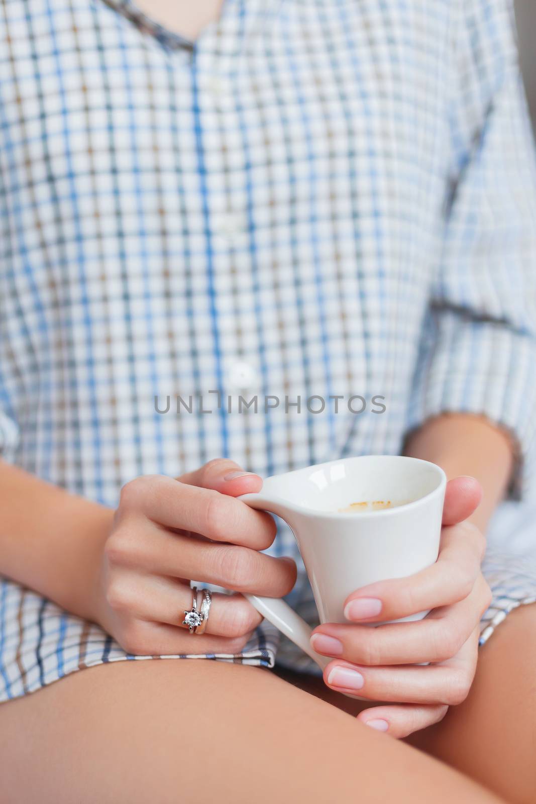 Woman in shirt is sitting on bed with a cup with hot coffee. Bright engagement and wedding rings sparkle on her finger.Cozy morning at home.