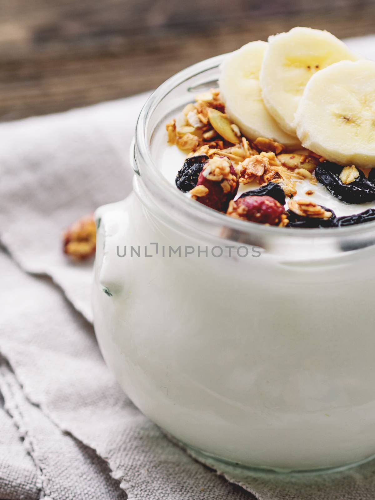 Natural homemade yogurt in a glass jar. Healthy food for breakfast with muesli. Jar with granola and banana slices on linen tablecloth on on wooden table.