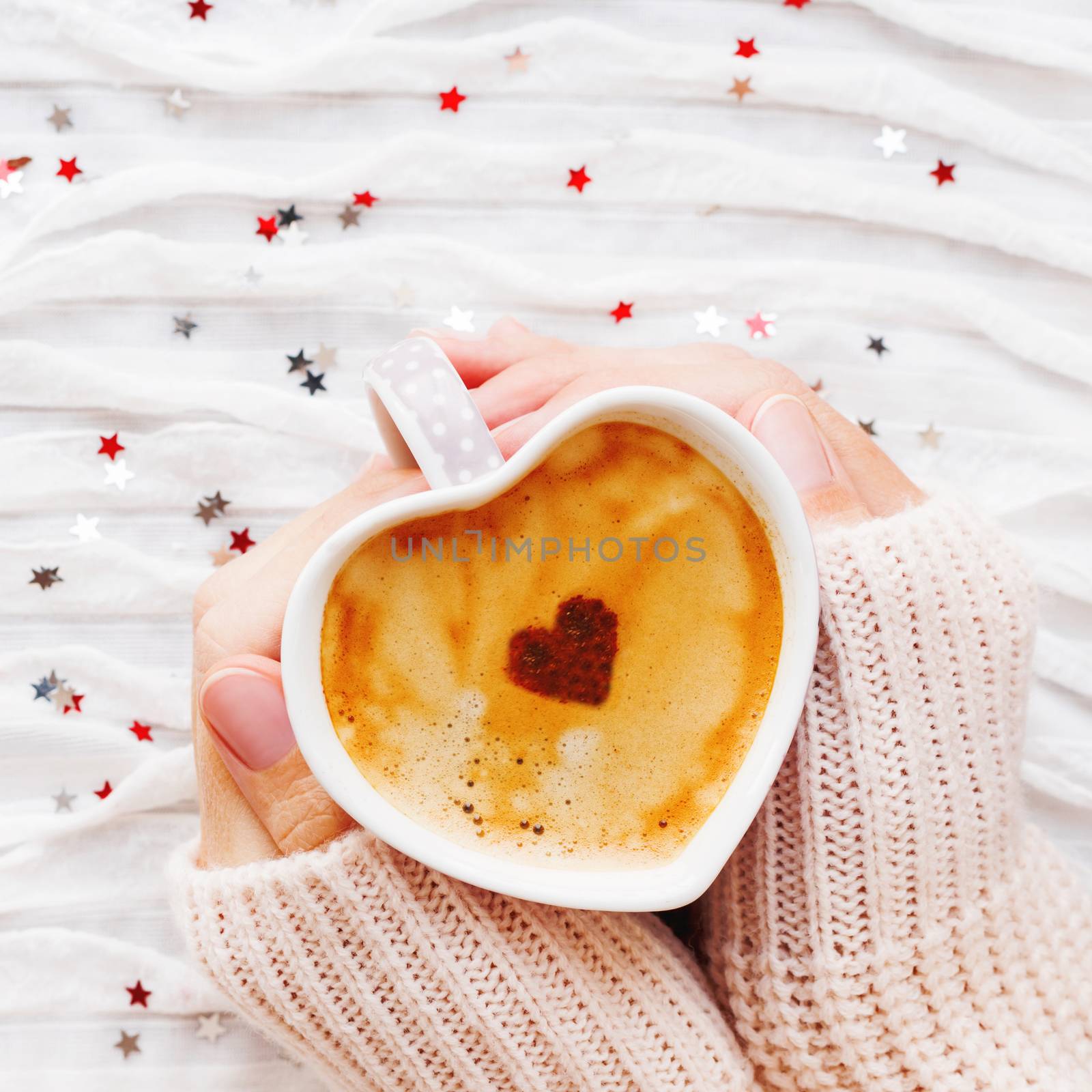 Woman holds a cup of hot coffee with cinnamon heart. Winter and Valentine's Day fabric background with sparkling silver and red confetti.