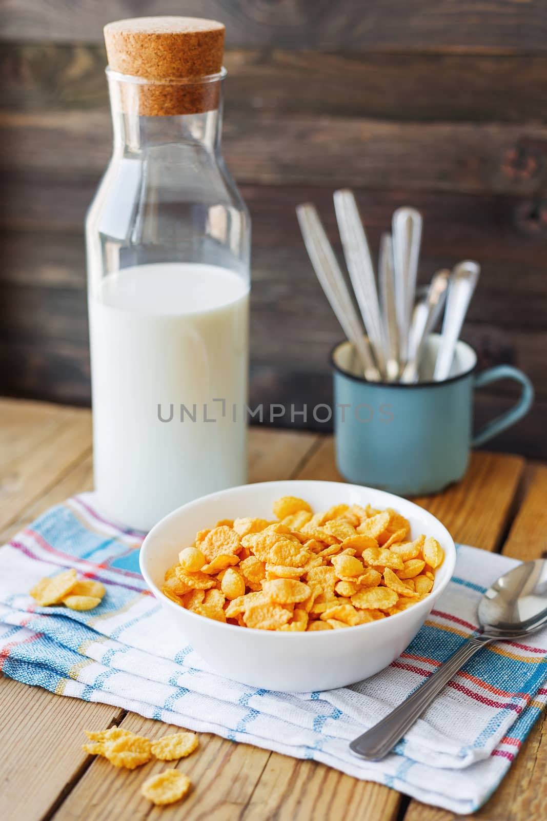Tasty corn flakes in white bowl with bottle of milk. Rustic wooden background with plaid napkin. Healthy crispy breakfast snack.