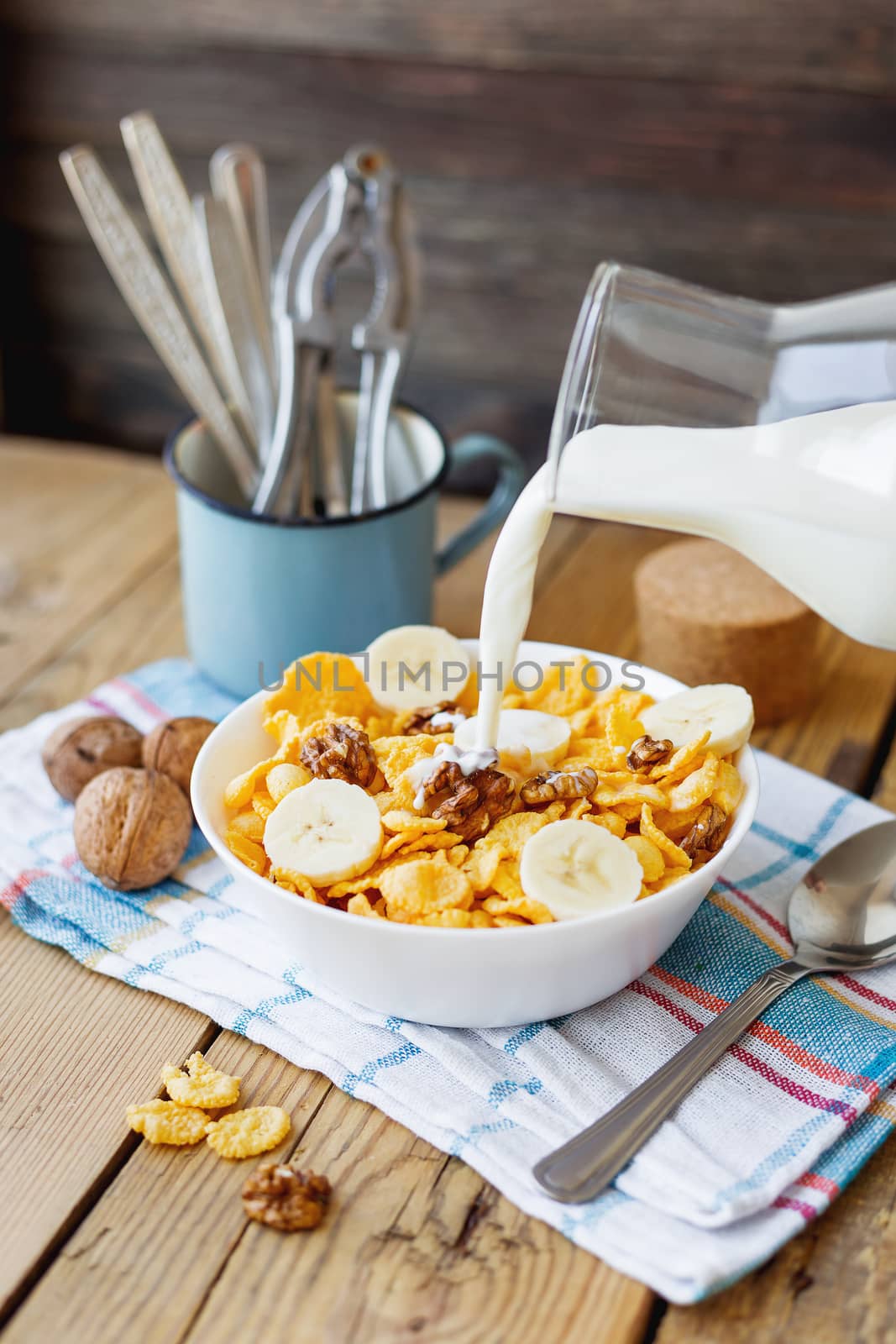 Milk from a bottle flows into the bowl with tasty corn flakes with walnuts and banana. Rustic wooden background with plaid napkin. Healthy crispy breakfast snack.