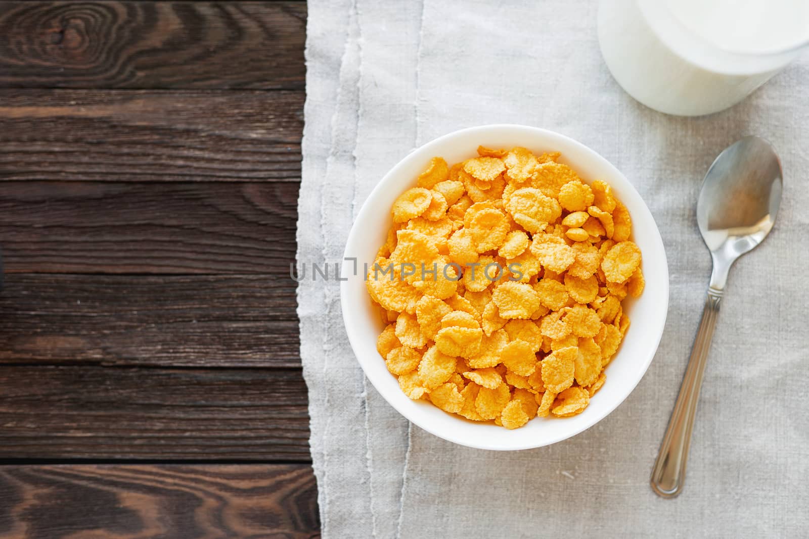 Tasty corn flakes in bowl with bottle of milk. Rustic wooden background with homespun napkin. Healthy crispy breakfast snack. Place for text. Top view, flat lay.