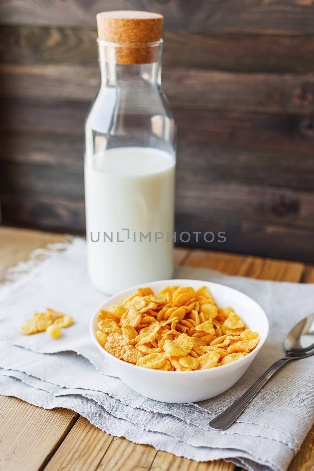 Tasty corn flakes in bowl with bottle of milk. Rustic wooden background with homespun napkin. Healthy crispy breakfast snack.