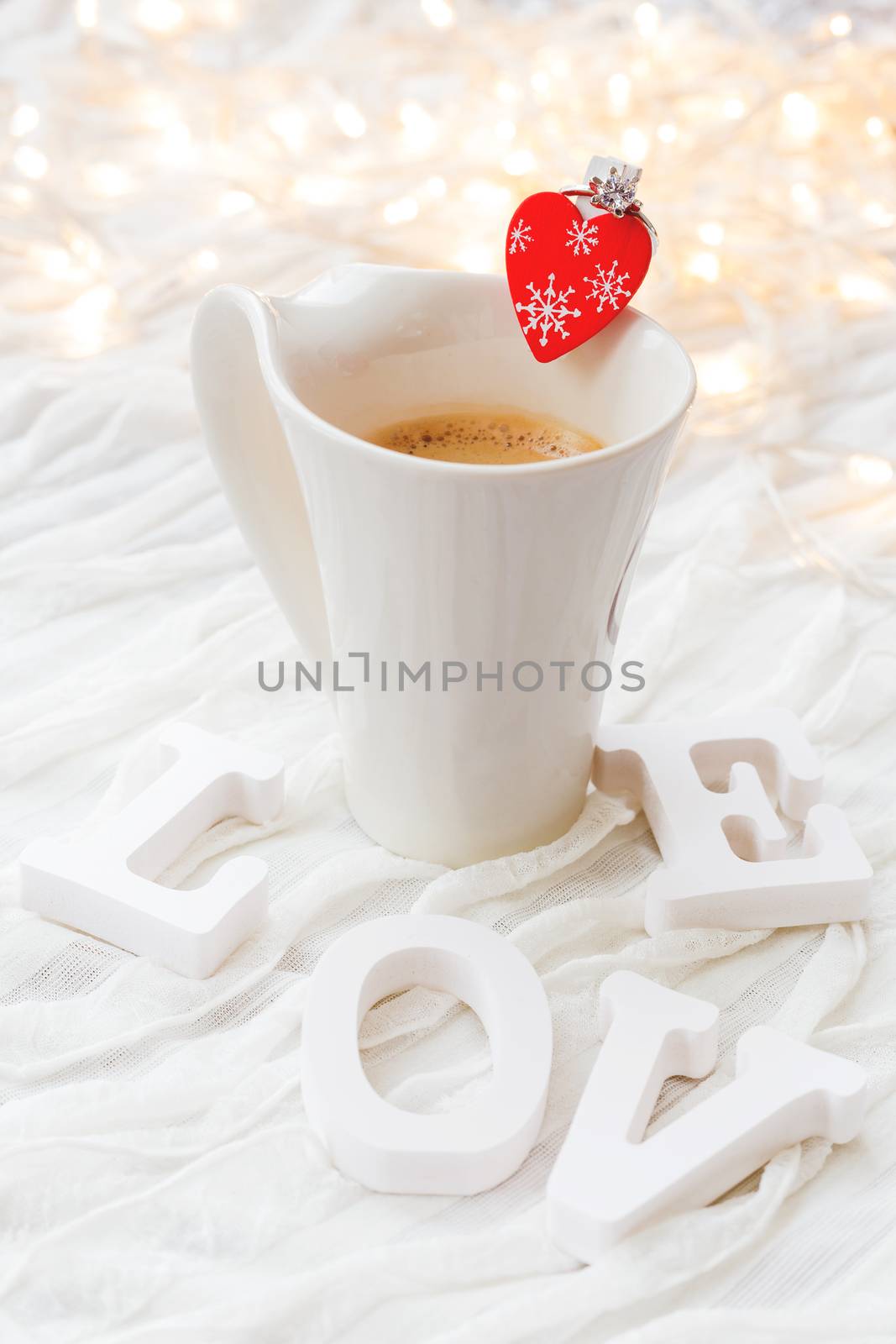 White cup of hot coffee with decorative heart and engagement diamond ring, symbol of love and marriage. Valentine's day background with light bulbs and decorations.